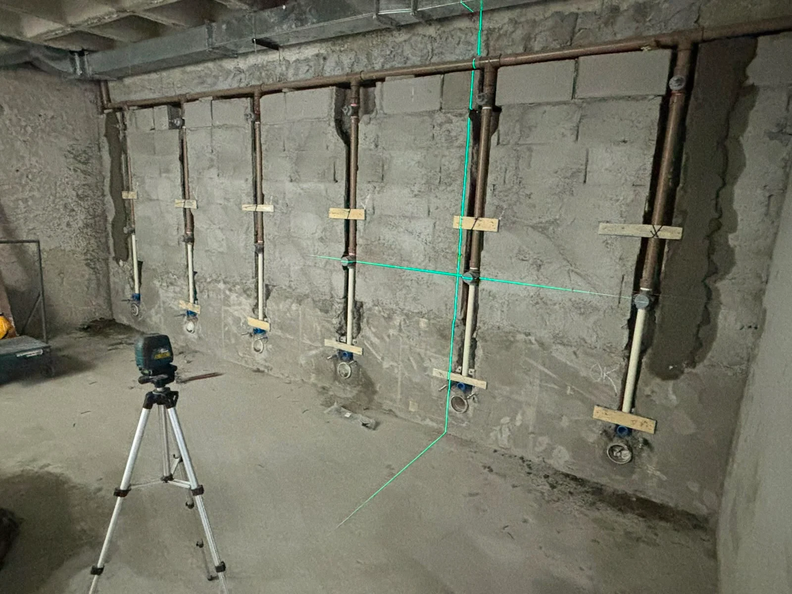 Tubulações embutidas em uma parede de alvenaria, com linhas verdes de nível a laser e um tripé à frente.