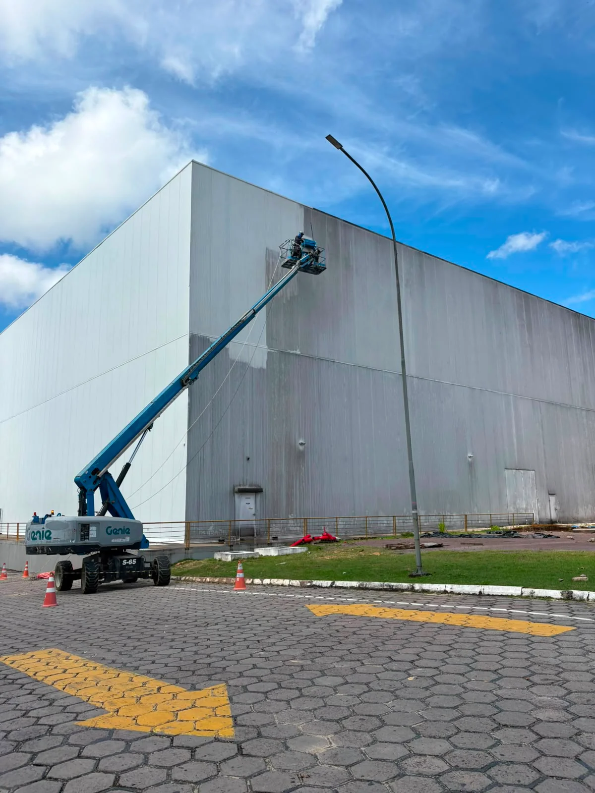Plataforma elevatória azul posicionada junto à fachada branca de um galpão, com um profissional no cesto.